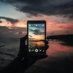 Mano che tiene un cellulare per scattare una foto con tramonto colorato sul mare.