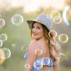 Una donna sorridente con un cappello grigio è circondata da tante foto bolle di sapone iridescenti in un prato verde.