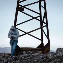 Uomo con fotocamera in mano, vicino a una struttura metallica arrugginita su una montagna. Il suo scatto fotografico significato cattura la vastità del paesaggio.