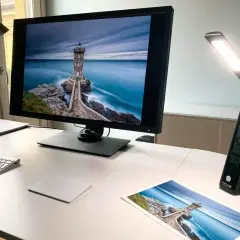 Scrivania con monitor che mostra un faro su scogli, una lampada da scrivania accesa e una stampa della stessa immagine. Luce inattinica.