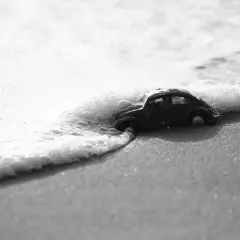 Un'auto giocattolo, in bianco e nero e colore, viene lambita dalle onde su una spiaggia sabbiosa.