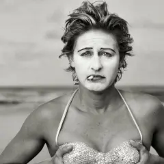 Annie Leibovitz foto: una donna con trucco da clown e sigaretta in bocca, in bikini scintillante, sulla spiaggia.