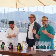 Three men stand behind a table with wine glasses and coffee bags, with a harbor in the background.