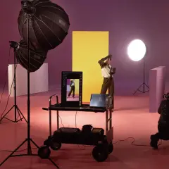 Nel set fotografico, una silhouette posa davanti a un pannello giallo. Luci e attrezzature creano un'atmosfera da studio.