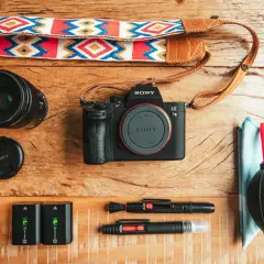 Un set da fotografia flat lay: fotocamera Sony, obiettivo, batterie, cinghia colorata e accessori per la pulizia.