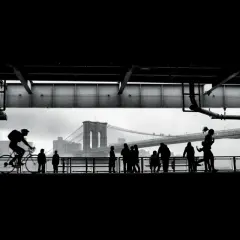 Ciclista in silhouette sul ponte di Brooklyn, con persone che passeggiano. Forse la foto più famosa del mondo, cattura un momento iconico.