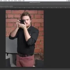Uomo con fotocamera in mano, sullo sfondo un muro di mattoni. Interfaccia di Photoshop aperta, con pannello "Proprietà" visibile, utile per la selezione rapida Photoshop.