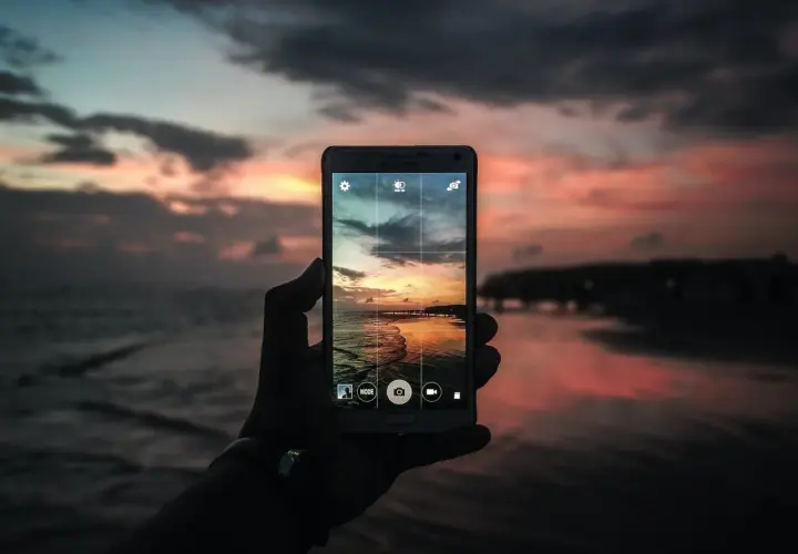 Mano che tiene un cellulare per scattare una foto con tramonto colorato sul mare.