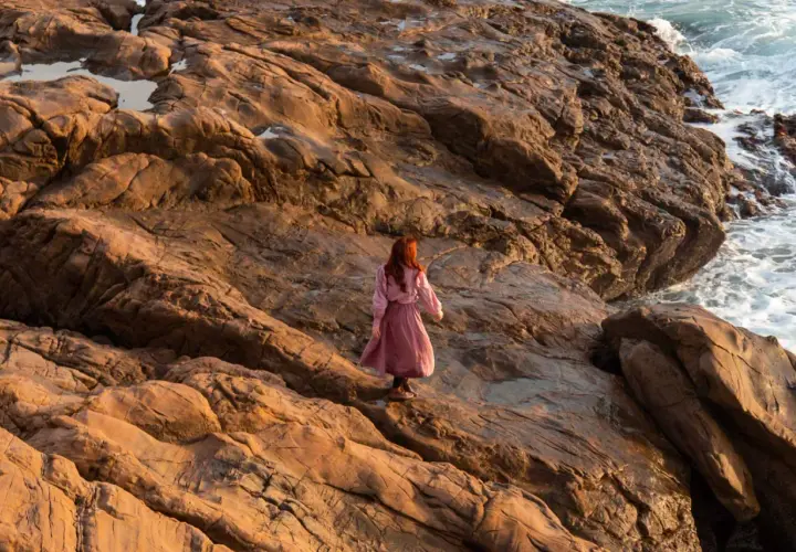Donna con capelli rossi cammina su rocce dorate al tramonto, evocando il significato del golden hour.