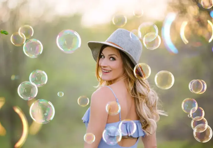 Una donna sorridente con un cappello grigio è circondata da tante foto bolle di sapone iridescenti in un prato verde.