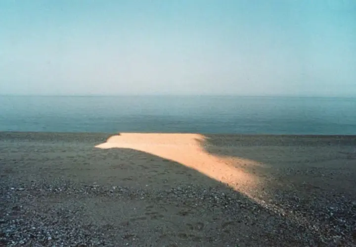 Una spiaggia con un'onda di luce dorata che attraversa la sabbia, tipica di una franco fontana foto. Il mare calmo e il cielo azzurro completano la scena.
