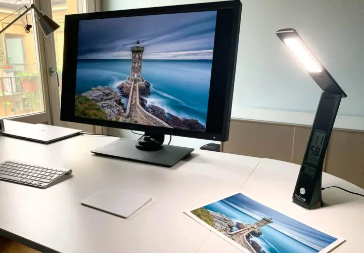 Scrivania con monitor che mostra un faro su scogli, una lampada da scrivania accesa e una stampa della stessa immagine. Luce inattinica.