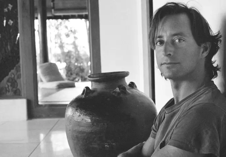 Ritratto di Herb Ritts accanto a un'antica giara, con uno scorcio di un salotto in stile tropicale sullo sfondo.