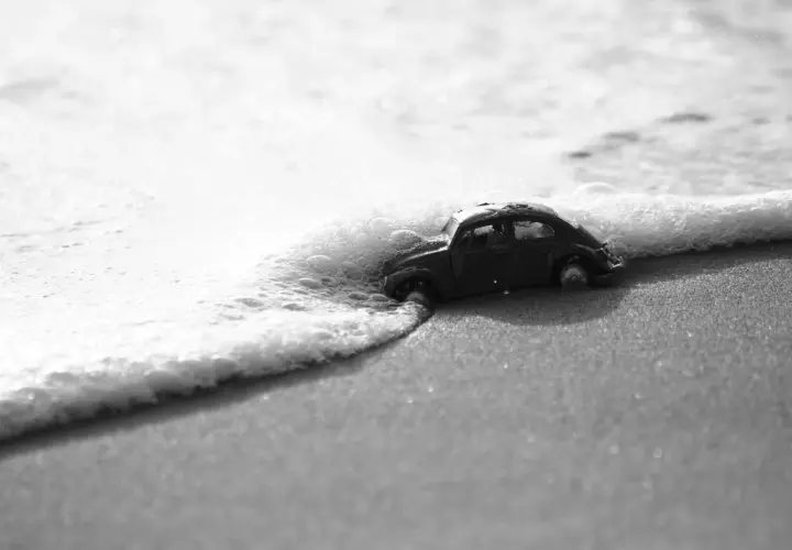 Un'auto giocattolo, in bianco e nero e colore, viene lambita dalle onde su una spiaggia sabbiosa.