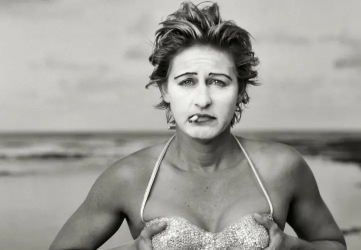 Annie Leibovitz foto: una donna con trucco da clown e sigaretta in bocca, in bikini scintillante, sulla spiaggia.