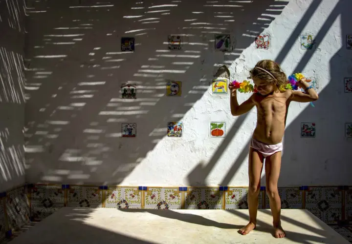 Bambina con collane di fiori, in piedi su un tavolo, sotto un gioco di luci e ombre. Le foto verticali sulla parete raccontano storie.