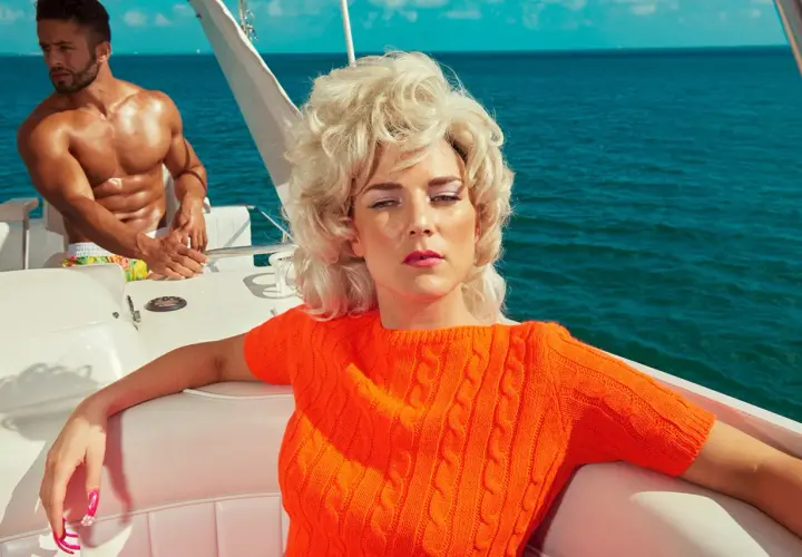 Kourtney Roy in un maglione arancione su una barca, con un uomo muscoloso sullo sfondo, in un'atmosfera estiva.