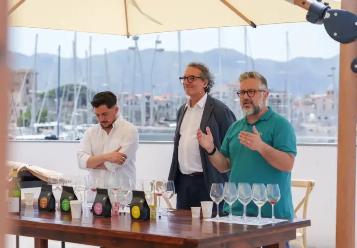 Three men stand behind a table with wine glasses and coffee bags, with a harbor in the background.