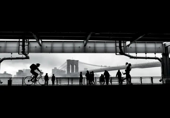 Ciclista in silhouette sul ponte di Brooklyn, con persone che passeggiano. Forse la foto più famosa del mondo, cattura un momento iconico.