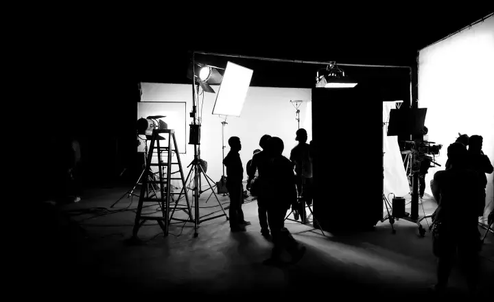 Scena di un set cinematografico con persone in silhouette, telecamere e un'intensa foto con luce.