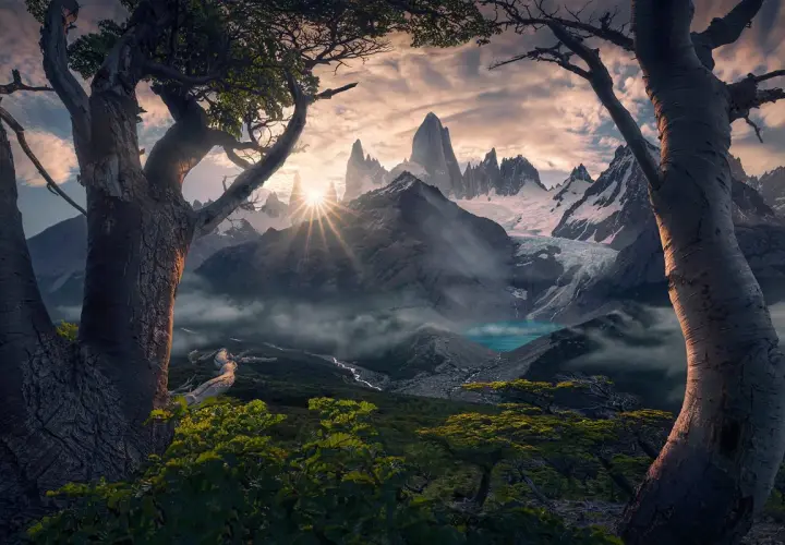Alba dorata sui picchi innevati del **max rive**, con un lago turchese e alberi antichi in primo piano.