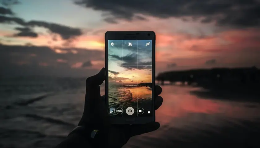 Mano che tiene un cellulare per scattare una foto con tramonto colorato sul mare.