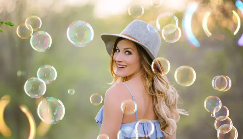 Una donna sorridente con un cappello grigio è circondata da tante foto bolle di sapone iridescenti in un prato verde.