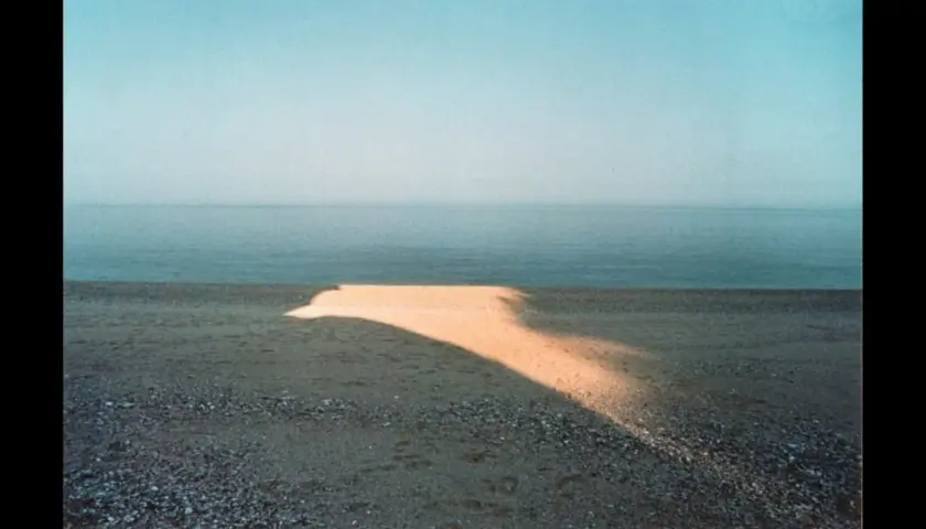 Una spiaggia con un'onda di luce dorata che attraversa la sabbia, tipica di una franco fontana foto. Il mare calmo e il cielo azzurro completano la scena.