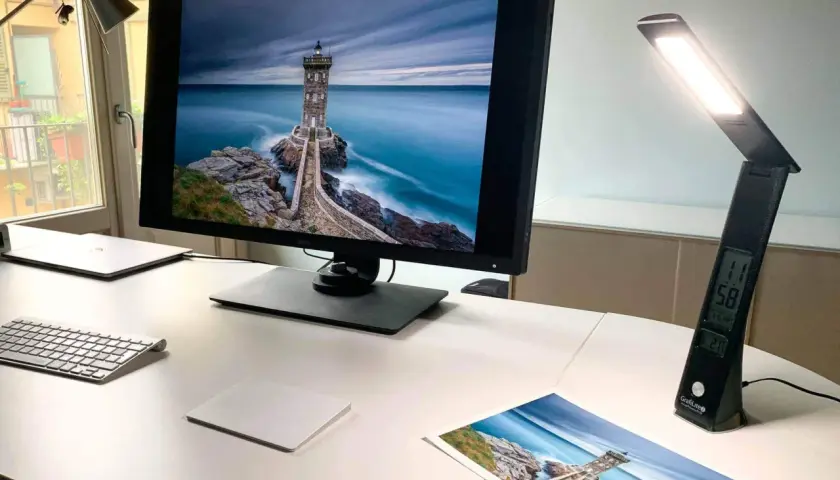Scrivania con monitor che mostra un faro su scogli, una lampada da scrivania accesa e una stampa della stessa immagine. Luce inattinica.