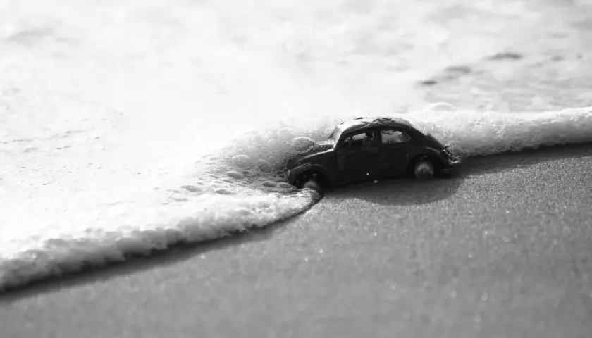 Un'auto giocattolo, in bianco e nero e colore, viene lambita dalle onde su una spiaggia sabbiosa.