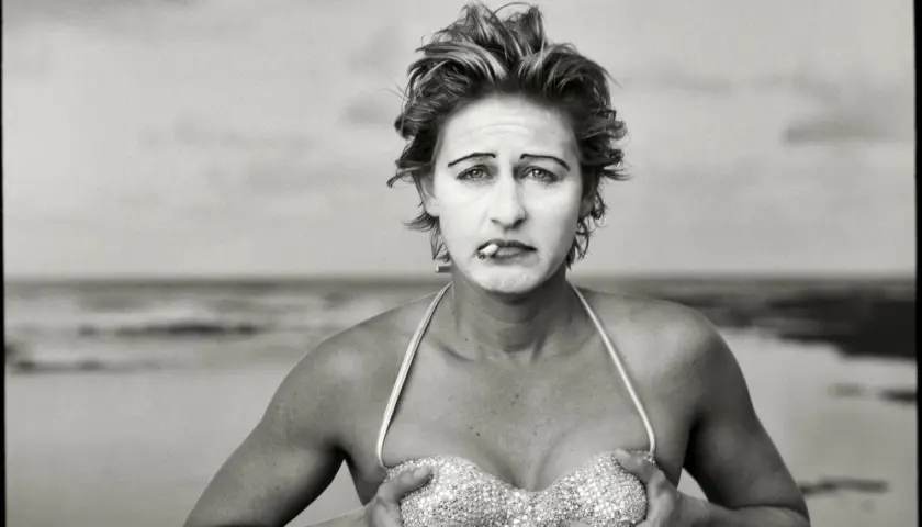 Annie Leibovitz foto: una donna con trucco da clown e sigaretta in bocca, in bikini scintillante, sulla spiaggia.