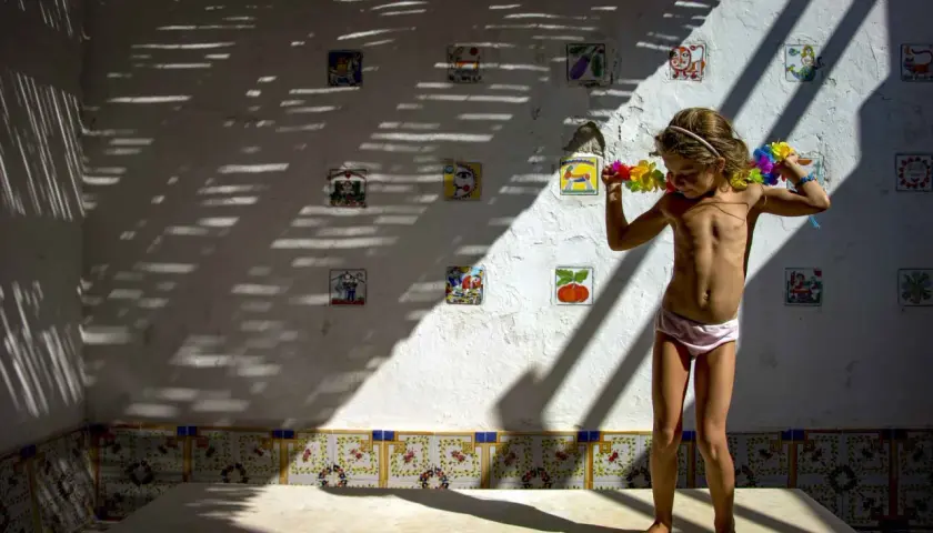 Bambina con collane di fiori, in piedi su un tavolo, sotto un gioco di luci e ombre. Le foto verticali sulla parete raccontano storie.