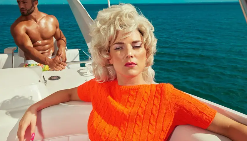 Kourtney Roy in un maglione arancione su una barca, con un uomo muscoloso sullo sfondo, in un'atmosfera estiva.