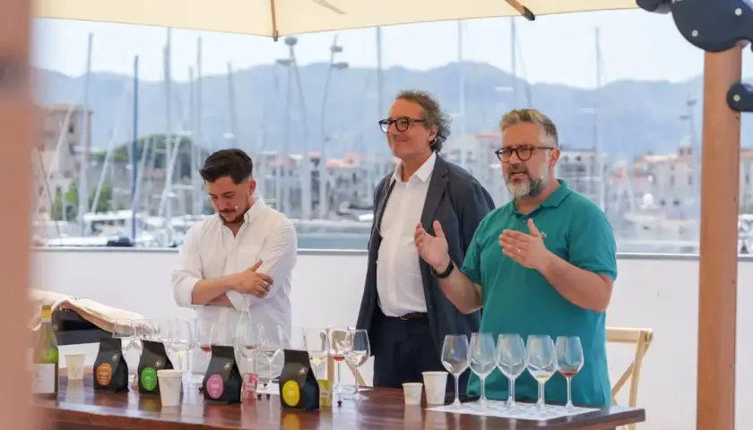 Three men stand behind a table with wine glasses and coffee bags, with a harbor in the background.