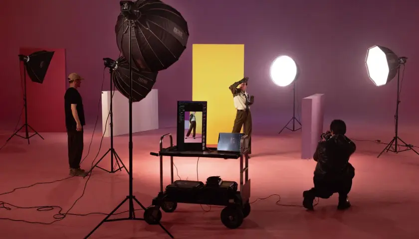 Nel set fotografico, una silhouette posa davanti a un pannello giallo. Luci e attrezzature creano un'atmosfera da studio.