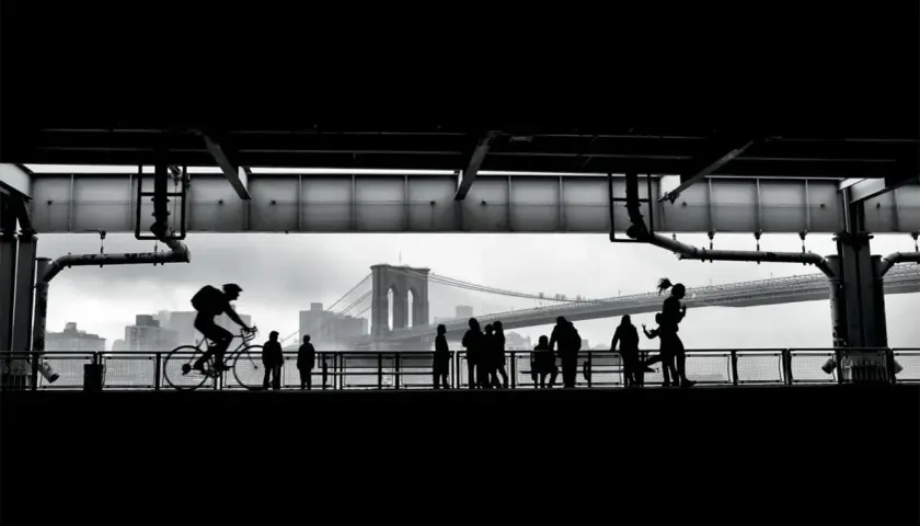 Ciclista in silhouette sul ponte di Brooklyn, con persone che passeggiano. Forse la foto più famosa del mondo, cattura un momento iconico.
