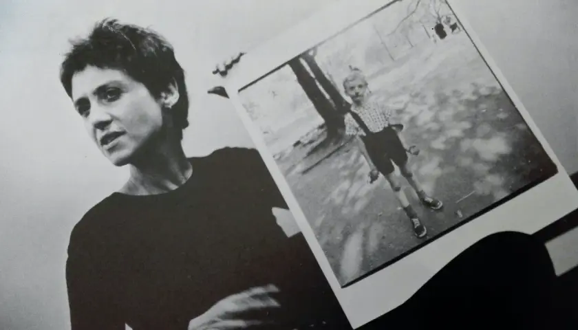 Diane Arbus, con la sua inconfondibile espressione, tiene in mano una delle sue celebri fotografie: un bambino in un parco.