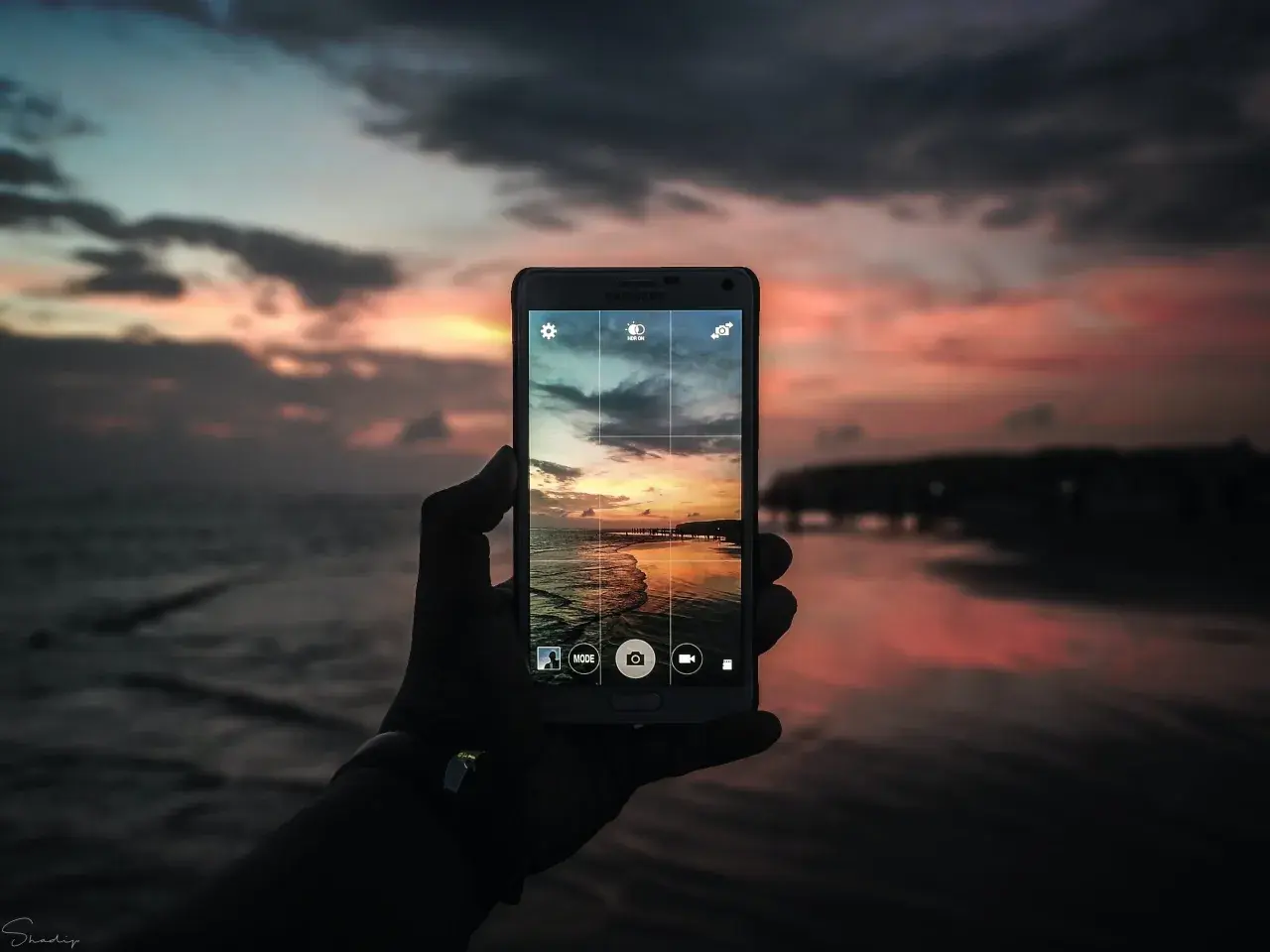 Mano che tiene un cellulare per scattare una foto con tramonto colorato sul mare.