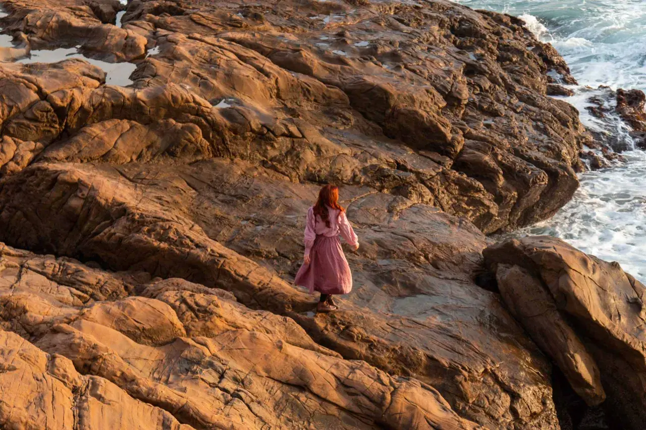 Donna con capelli rossi cammina su rocce dorate al tramonto, evocando il significato del golden hour.