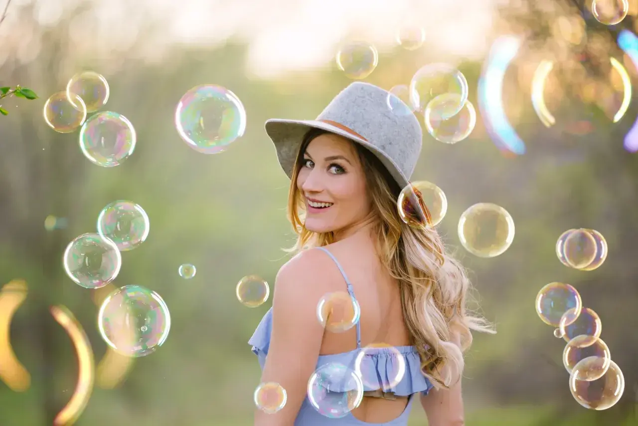 Una donna sorridente con un cappello grigio è circondata da tante foto bolle di sapone iridescenti in un prato verde.