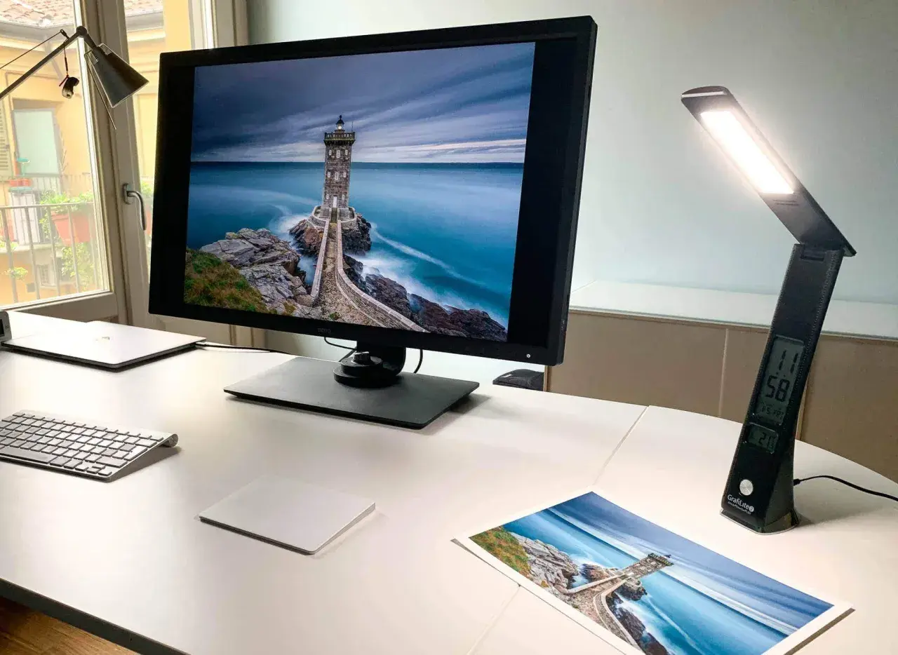 Scrivania con monitor che mostra un faro su scogli, una lampada da scrivania accesa e una stampa della stessa immagine. Luce inattinica.