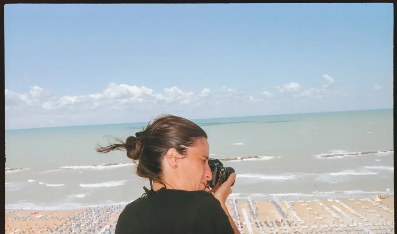 Federica Valabrega cattura la bellezza del mare con la sua macchina fotografica, mentre una spiaggia affollata si estende sotto un cielo azzurro.