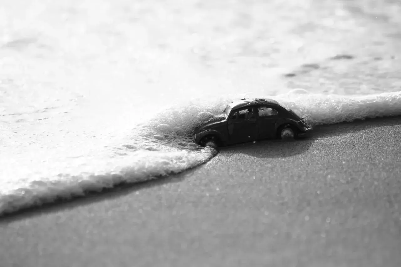 Un'auto giocattolo, in bianco e nero e colore, viene lambita dalle onde su una spiaggia sabbiosa.