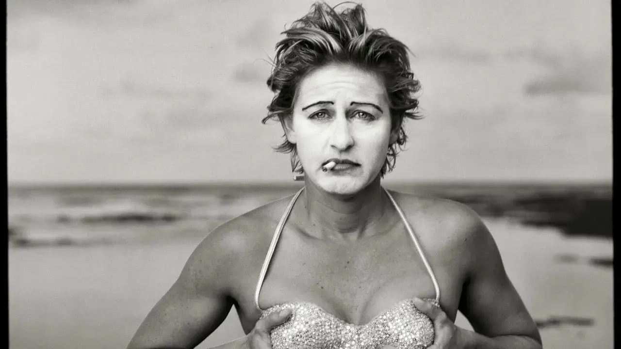 Annie Leibovitz foto: una donna con trucco da clown e sigaretta in bocca, in bikini scintillante, sulla spiaggia.