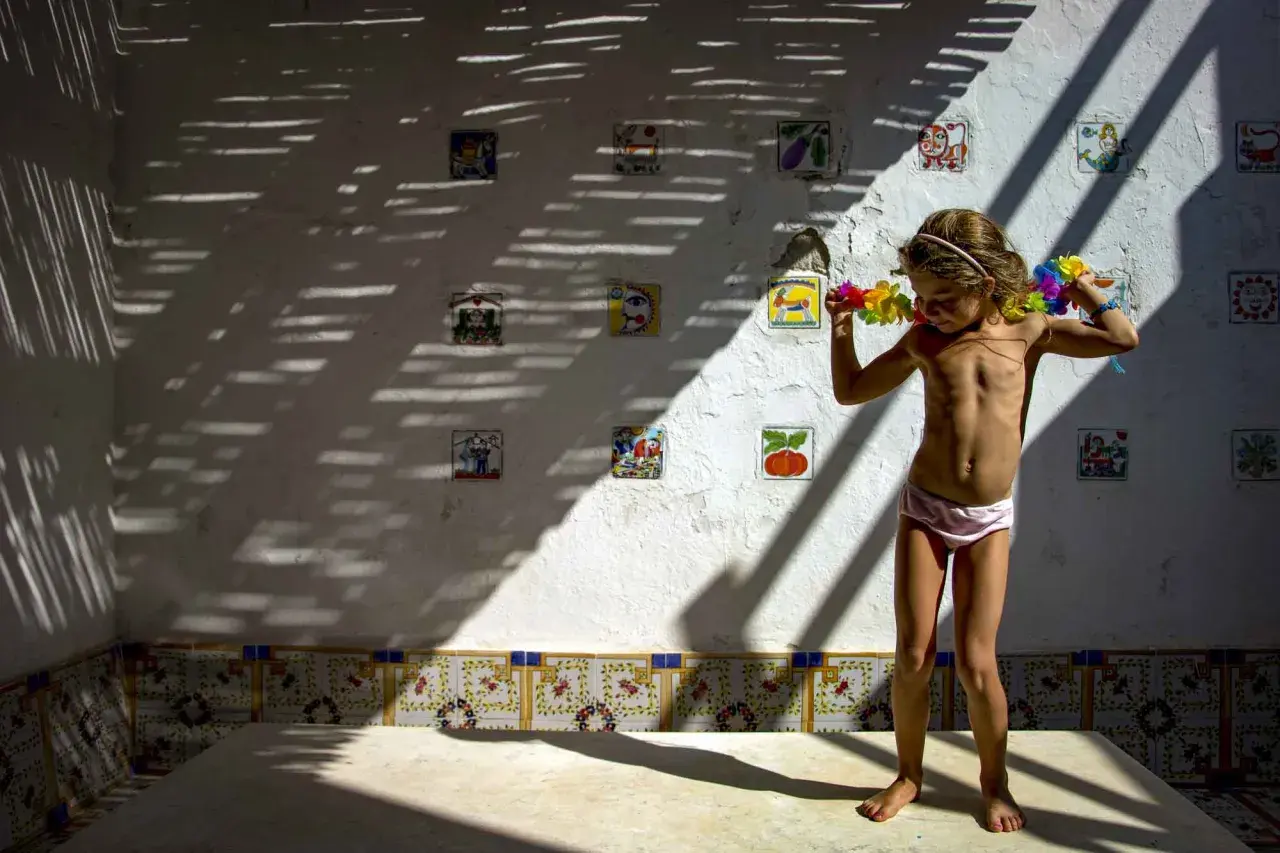 Bambina con collane di fiori, in piedi su un tavolo, sotto un gioco di luci e ombre. Le foto verticali sulla parete raccontano storie.