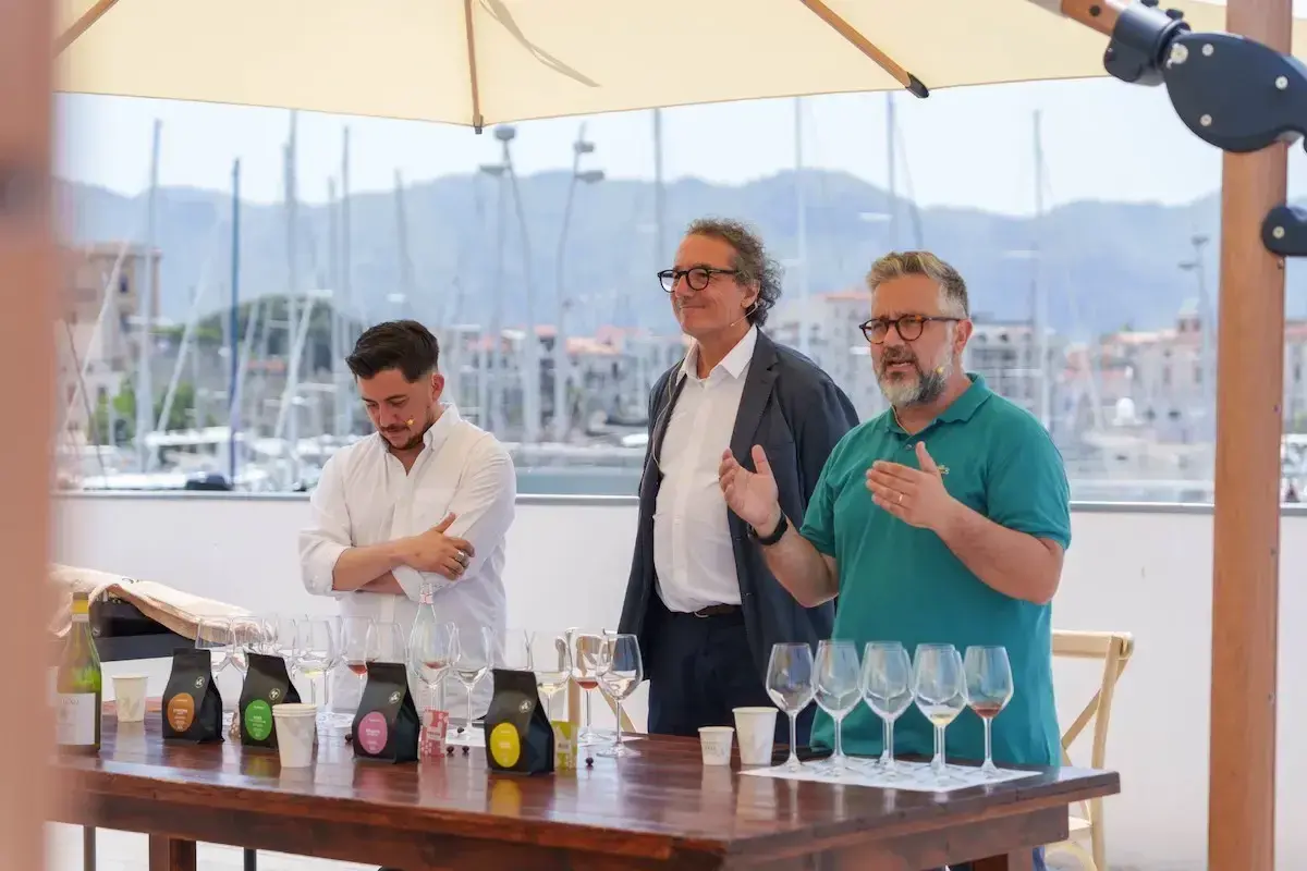 Three men stand behind a table with wine glasses and coffee bags, with a harbor in the background.