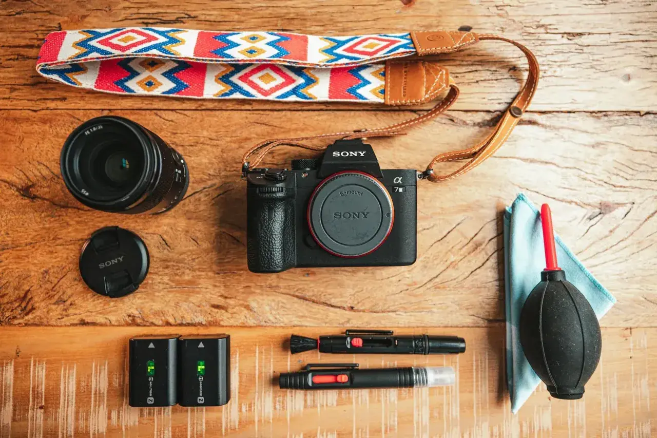 Un set da fotografia flat lay: fotocamera Sony, obiettivo, batterie, cinghia colorata e accessori per la pulizia.