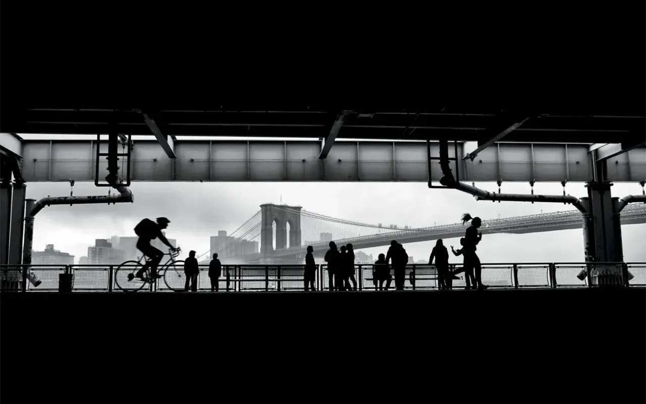 Ciclista in silhouette sul ponte di Brooklyn, con persone che passeggiano. Forse la foto più famosa del mondo, cattura un momento iconico.