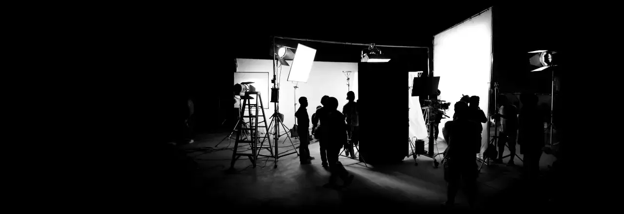 Scena di un set cinematografico con persone in silhouette, telecamere e un'intensa foto con luce.