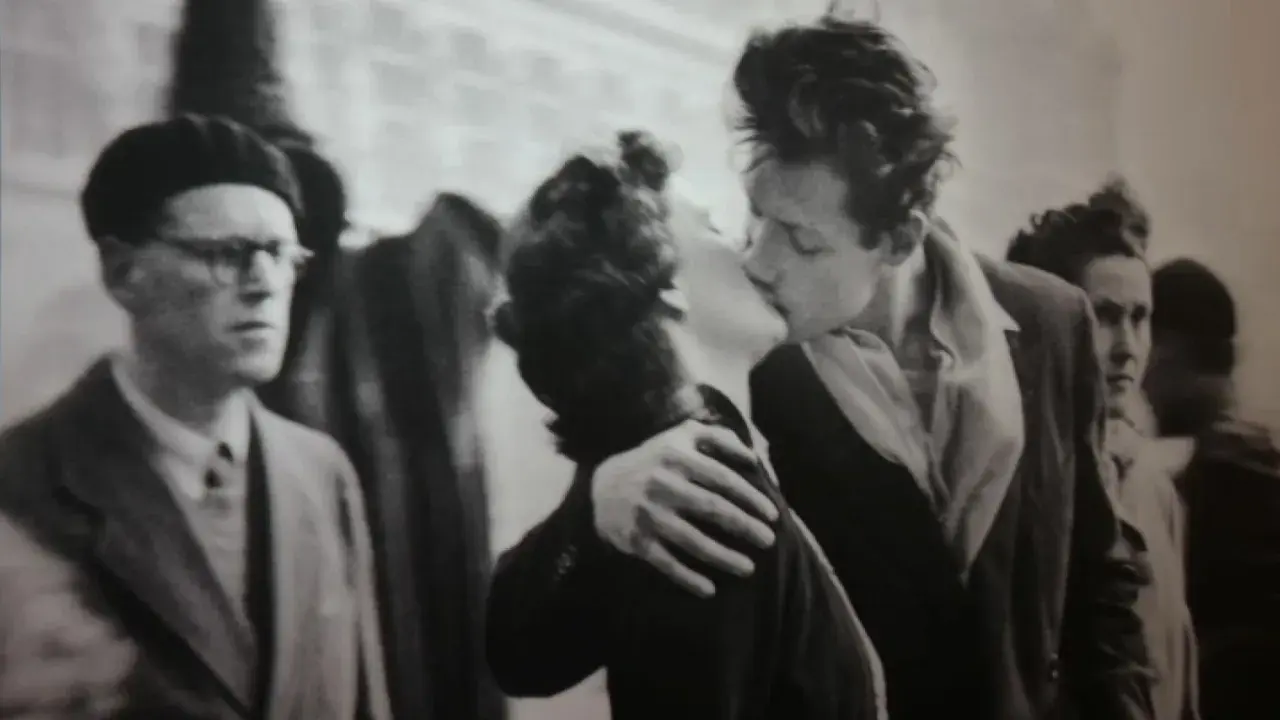 Un bacio appassionato catturato in uno degli iconici robert doisneau opere. Un uomo con berretto e occhiali osserva la scena.